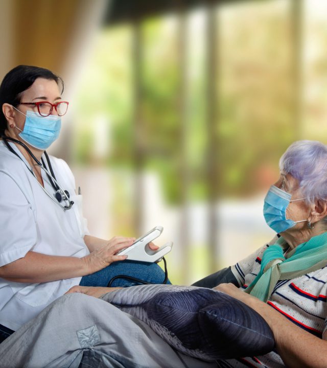 A geriatric doctor visiting an elderly patient at home to take blood pressure. However, both the patient and the geriatrician doctor are required to wear masks during a pandemic.
